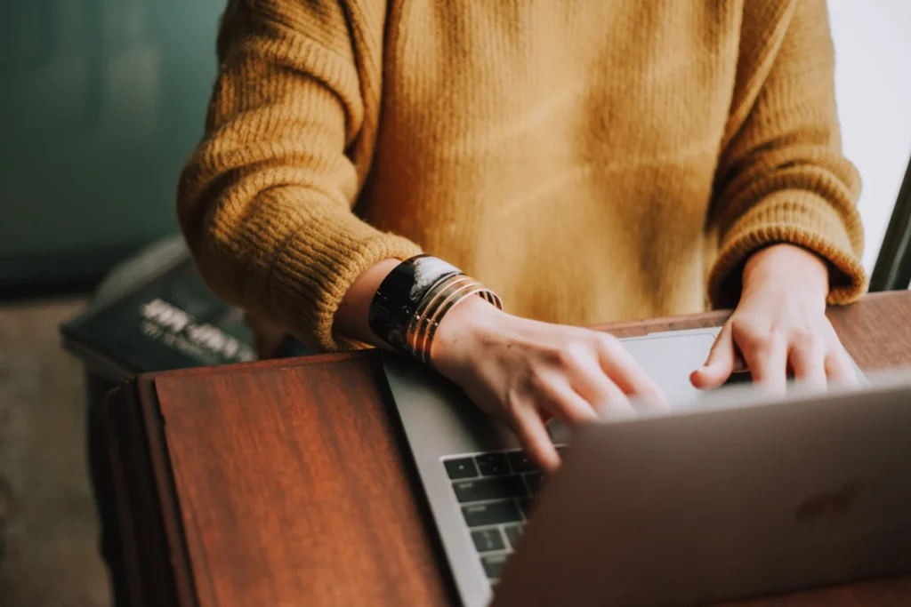 Person typing on a laptop wearing a mustard sweater and bracelets.