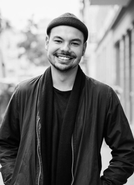Smiling man wearing a hat and jacket in an outdoor urban setting.